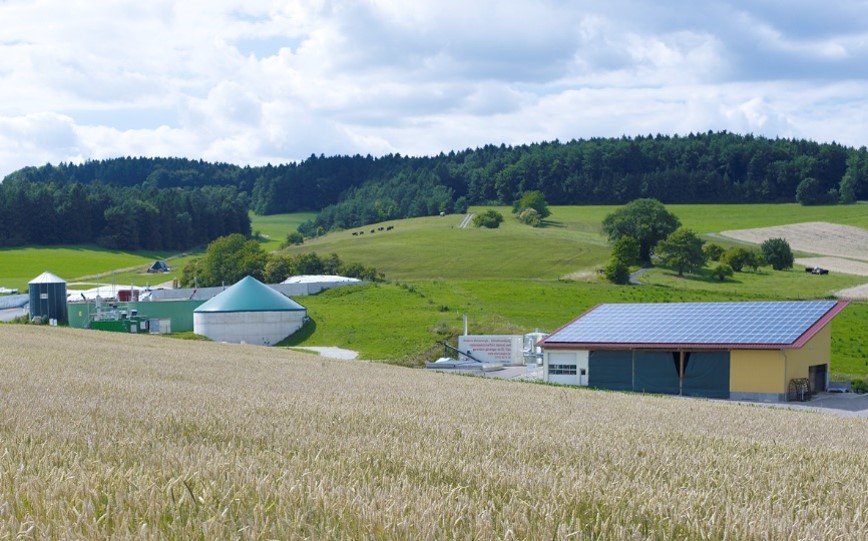 Das Bild zeigt die Energie- und Biogasanlage des Bioenergiedorfs Mauenheim. Die Anlage und Gebäude stehen in ländlicher Umgebung. Im Vordergrund ein Felder, im Hintergrund ein Wald vor wolkigem Himmel.