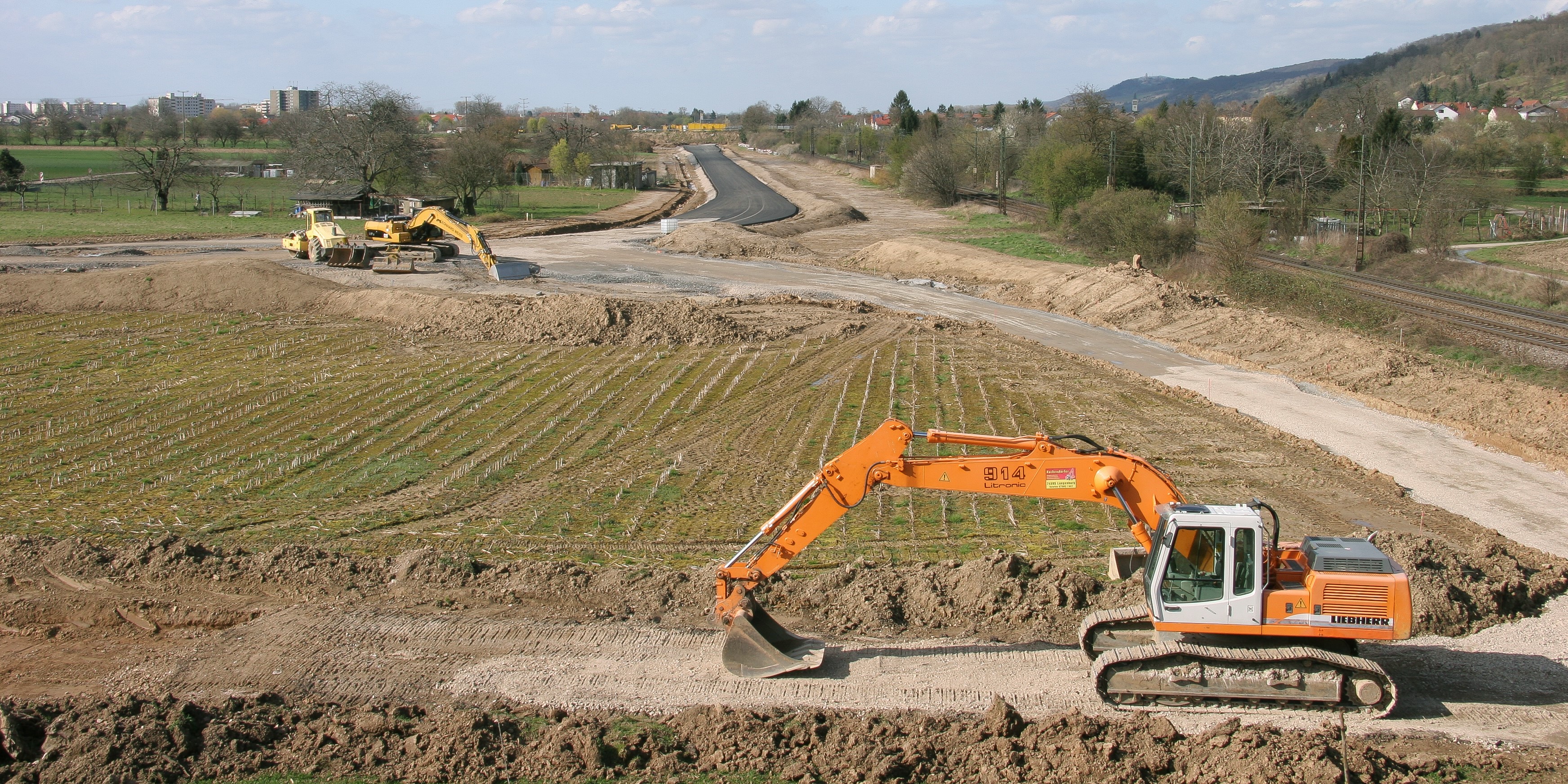 Fortführung einer neuen, bereits begonnenen Straße durch Felder und Wiesen. Im Vordergrund bereitet ein Bagger den Boden für weitere Baumaßnahmen vor.