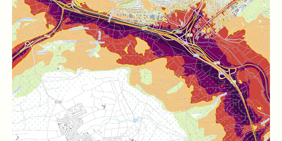 Ausschnitt aus der Lärmkartierung 2022. Man sieht eine Landkarte, auf der eine Autobahn an einer Siedlung vorbeiführt. Entlang der vielbefahrenen Straße zeichnet sich deutlich der von ihnen ausgehende „Lärmteppich“ ab. Die Lärmschutzwand entlang der Autobahn reduziert den Verkehrslärm im Siedlungsbereich.]