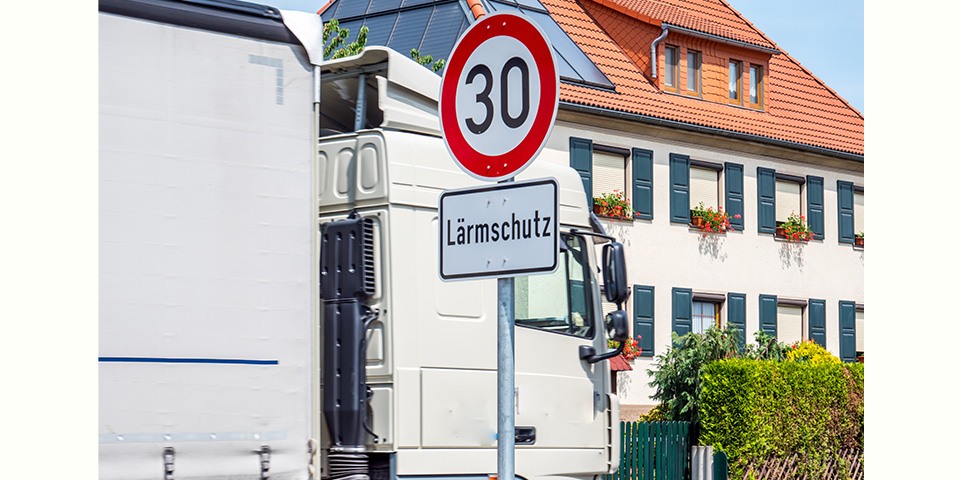 Verkehrszeichen „Höchstgeschwindigkeit 30 km/h“ mit dem Zusatzschild „Lärmschutz“.Dahinter ein vorbeifahrender Lastwagen, im Hintergund ein Wohnhaus.