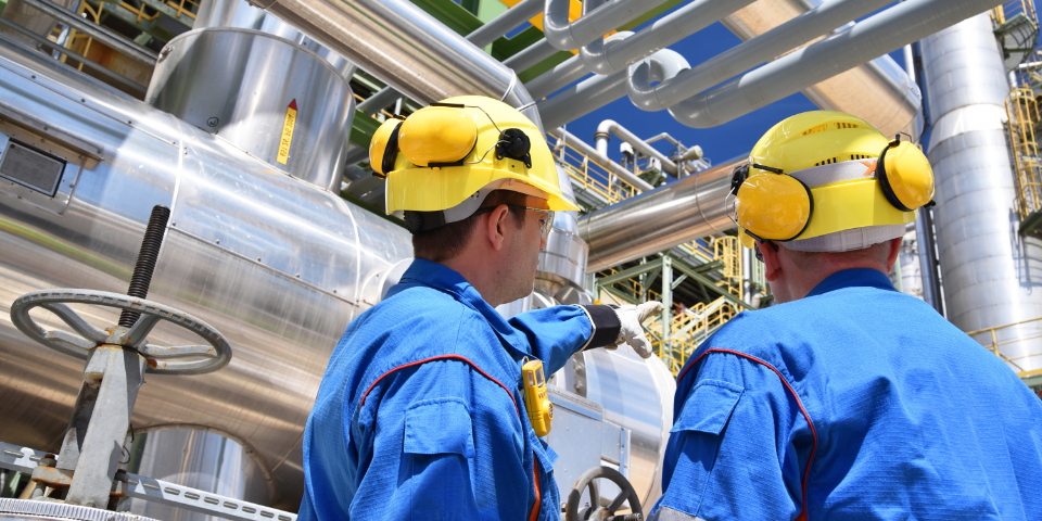 Zwei Personen in Arbeitskleidung und mit Schutzhelm vor einer Industrieanlage.