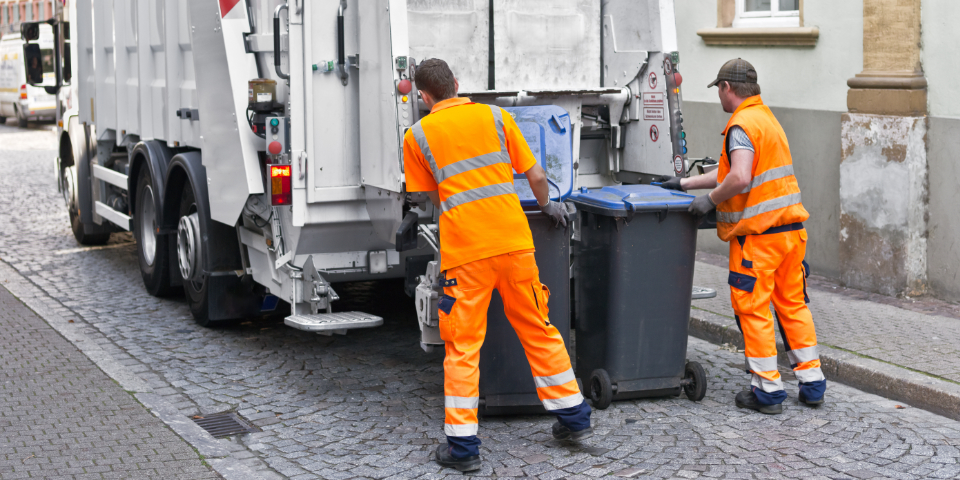 Zwei Mitarbeiter eines Abfallwirtschaftsbetriebes schieben Mülltonnen an ein Müllauto zum Entleeren.